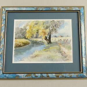 May include: Watercolor landscape painting of a river scene with a tree in the center. The painting is framed in a blue and gold decorative frame. The artwork depicts a serene outdoor scene with a river, trees, and a small building in the distance.