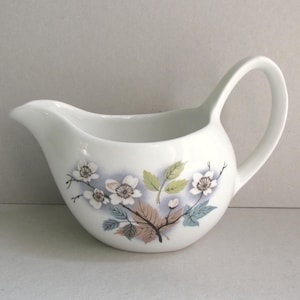May include: A white ceramic creamer pitcher featuring a curved handle and a pouring spout. The exterior is decorated with a botanical illustration of white flowers, brown and green leaves, and soft blue shading on the rounded body of the vessel.