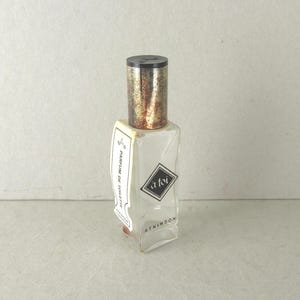 May include: A clear, rectangular perfume bottle with a black cap and a label that reads "a foi" in a diamond shape. The bottle has a gold and brown speckled top and a white label that reads "PARFUM DE TOILETTE".