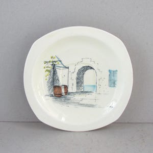 May include: A white ceramic plate featuring a hand-drawn illustration of an arched doorway, barrels, and a window. The artwork uses blue, green, and brown tones. The plate has a slightly irregular, scalloped edge.