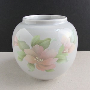 May include: A white ceramic vase with a floral design. The vase has a round body and a wide mouth. The flowers are pink and green and are painted on the vase.