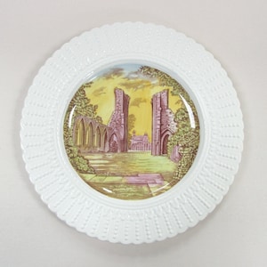 May include: White decorative plate with a scalloped edge. The center features a colorful illustration of a historic building ruin with a yellow and green landscape. The plate is round.