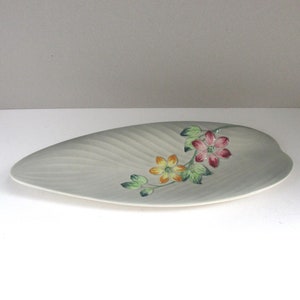 May include: A leaf-shaped ceramic serving dish in a pale grey colour with a subtle ribbed texture. The dish features a hand-painted floral motif with two blossoms in shades of yellow and pink, accompanied by green leaves along a thin brown stem.