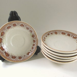 May include: Six off-white ceramic saucers with a brown speckled border. One saucer is on a black stand, the others stacked. The saucers have a simple, classic design, suitable for various table settings. Each saucer has a diameter of approximately 15 cm.