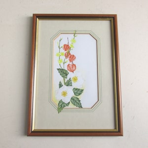 May include: Framed watercolor painting featuring white flowers with yellow centers and green leaves, alongside orange and yellow berries. The artwork is set against a cream background within a brown wooden frame with a gold inner border. The painting is a botanical illustration.