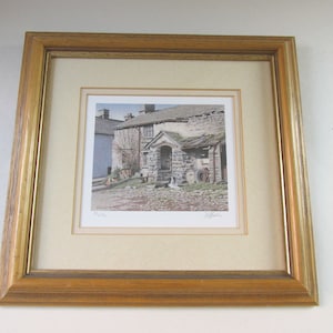May include: Framed print of a stone cottage with a slate roof, set in a rural landscape. The artwork is displayed in a gold-toned wooden frame with a cream-colored mat. The print features a detailed depiction of the building and its surroundings.