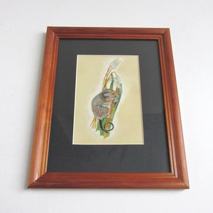 May include: A framed watercolor painting of a small brown mouse climbing a stalk of green grass with white flowers. The painting is set against a cream background and framed in a dark wood frame.