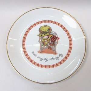 May include: White ceramic plate with a gold rim featuring a cartoon illustration of two figures in a wicker chair. The plate has a red and yellow plaid border and the text "Make every day a happy day".