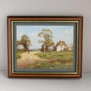May include: Framed landscape print featuring a dirt road leading to a white cottage with a red roof. The artwork depicts a serene countryside scene with trees, a small pond, and a blue sky. The frame is brown with a gold inner border.