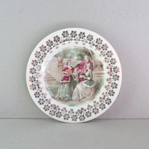 May include: White decorative plate with a floral border in brown and gold. The center features a scene of three figures in period clothing, with one playing a stringed instrument. The plate has a vintage aesthetic.