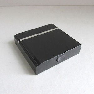 May include: A black rectangular box with a silver stripe and a small decorative element on the top. The box has a hinged lid and appears to be made of a solid, dark material. The design is simple and elegant.