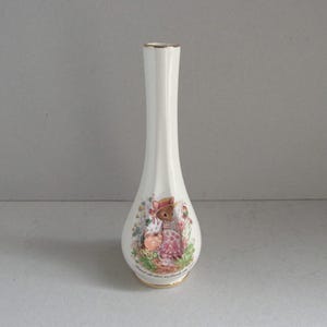 May include: A tall, cream-coloured ceramic vase with a gold rim. The vase has a painted illustration of a mouse in a pink dress and hat, holding a basket of flowers. The design is surrounded by floral elements.