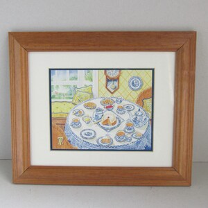 May include: Framed watercolour painting depicting a tea party. A round table is set with a blue and white patterned tablecloth, crockery, and cake. The painting is in a wooden frame with a white mount.