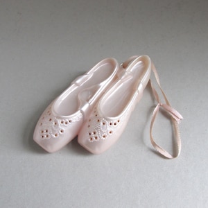 May include: A pair of pale pink ballet slippers with decorative cutouts on the toe. The satin slippers have matching ribbons attached. The shoes are displayed on a light grey surface.