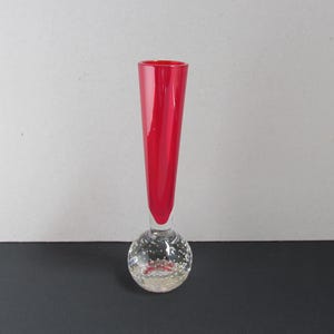 May include: A tall, slender red glass vase with a clear, bubble-filled spherical base. The vase is approximately 8 inches tall, with a vibrant red color extending from the top to the base's connection point. The base is clear with small bubbles.