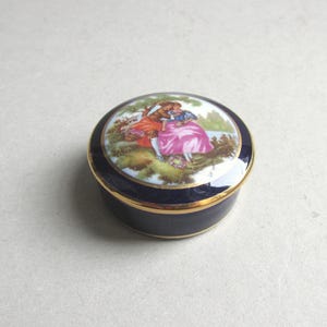 May include: A small, round trinket box with a dark blue base and a gold rim. The lid features a painted scene of two figures in a garden setting, with a pink dress and a blue dress. The box is likely made of porcelain.