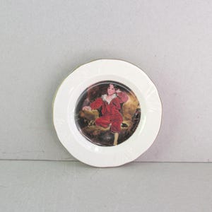 May include: White decorative plate with a gold rim. The center features a painting of a person in a red outfit. The plate has a scalloped edge design. A classic decorative item.