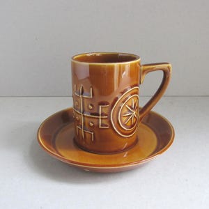 May include: A brown ceramic mug and saucer set. The mug has a handle and is decorated with a geometric pattern and a starburst design. The saucer is round and matches the mug's color and design.
