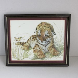 May include: Framed watercolor print of a tiger cub lying in grass with a hummingbird. The tiger cub has brown and black stripes and is framed in a dark brown frame with a burgundy inner border. The artwork is signed.