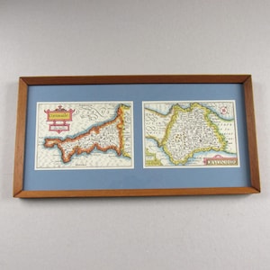May include: Framed vintage maps of Cornwall and Devonshire, England. The maps are in a wooden frame with a light blue matting. The maps are detailed with colorful illustrations and text, showcasing the historical cartography of the regions.