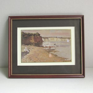May include: A framed landscape painting depicting a coastal scene with a sandy beach, rocky cliffs, and a small boat on the water. The artwork is enclosed in a dark reddish-brown wooden frame with a dark brown mat and a thin gold inner border.