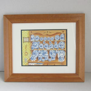 May include: A framed print depicting a kitchen dresser displaying blue and white china. The artwork shows plates, teacups, and teapots. The frame is made of light brown wood. A clock and ladle are also visible.