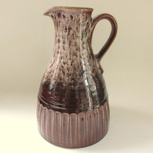 May include: A tall, brown ceramic jug with a handle and spout. The jug has a textured surface with vertical grooves at the base and a mottled brown and white pattern on the upper portion. A decorative piece for the home.