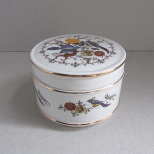 May include: A white ceramic trinket box with a lid, adorned with a floral and bird design in blue, yellow, and brown. The box has gold trim and a round shape, suitable for storing small items.