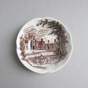 May include: A white, scalloped-edge plate with a brown and white transferware design. The scene depicts a castle with a horse-drawn carriage, trees, and figures. The plate has a diameter of approximately 6 inches.
