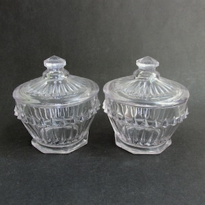May include: Two clear, lidded glass jars with a faceted design. The jars have a hexagonal base and a decorative, ridged lid with a diamond-shaped finial. The jars are displayed on a dark surface.