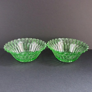 May include: Two green glass bowls with a scalloped rim. The bowls have a textured, patterned design on the exterior and a smooth interior. The bowls are made of a transparent green glass.