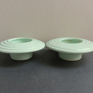 May include: Two light green ceramic candle holders with a wide, flat base and a ring design around the top.