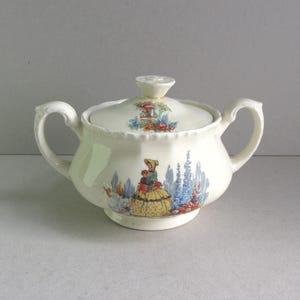 May include: Cream-colored ceramic sugar bowl with lid and handles. The bowl features a colorful illustration of a woman in a yellow dress and a garden scene. The lid has a small knob and a floral design.