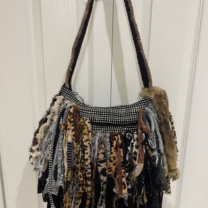 Puede incluir: Un bolso de hombro de crochet negro y marr&oacute;n con un detalle de flecos. Los flecos est&aacute;n hechos de varias telas, incluyendo estampado de leopardo, negro y gris. El bolso tiene un frente brillante cubierto de diamantes de imitaci&oacute;n.