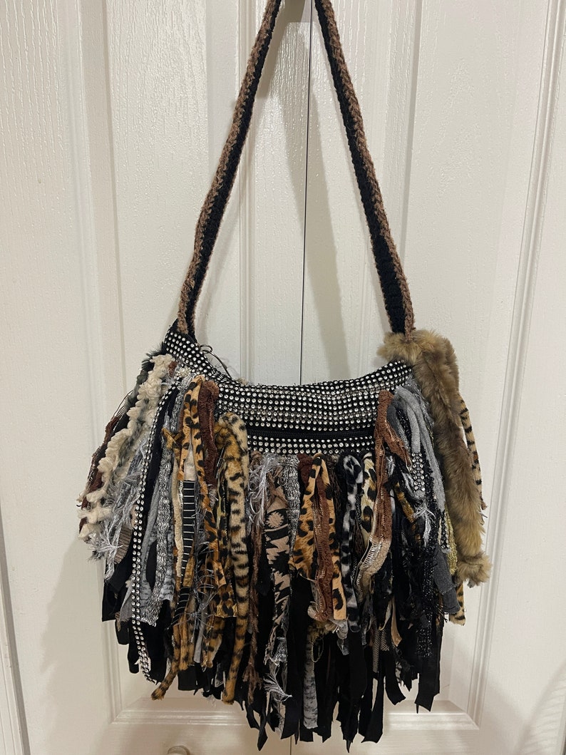 Puede incluir: Un bolso de hombro de crochet negro y marr&oacute;n con un detalle de flecos. Los flecos est&aacute;n hechos de varias telas, incluyendo estampado de leopardo, negro y gris. El bolso tiene un frente brillante cubierto de diamantes de imitaci&oacute;n.