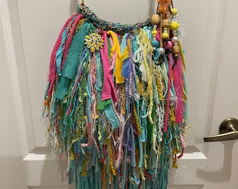 May include: A colourful crocheted bag with a long fringe and beaded strap. The bag is made of various colours of yarn and fabric, including blue, green, yellow, pink, and purple.