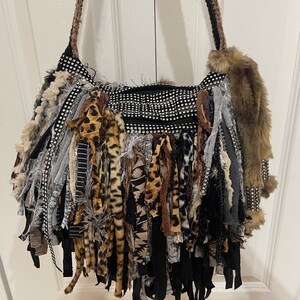 Puede incluir: Un bolso negro con una larga franja hecha de varias telas, incluyendo estampado de leopardo, rayas blancas y negras y gris. El bolso tiene una correa de cadena plateada y est&aacute; decorado con diamantes de imitaci&oacute;n.