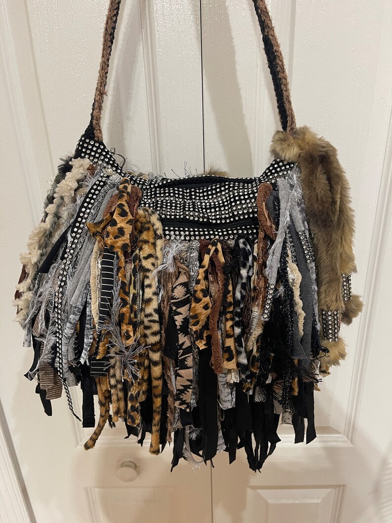 Puede incluir: Un bolso negro con una larga franja hecha de varias telas, incluyendo estampado de leopardo, rayas blancas y negras y gris. El bolso tiene una correa de cadena plateada y est&aacute; decorado con diamantes de imitaci&oacute;n.