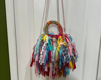 May include: A vibrant, handmade handbag with a circular form and a long strap. The bag is adorned with fabric strips in diverse hues, such as pink, blue, yellow, and white. The handle is wrapped in coordinating fabric. The bag is suspended on a white door.