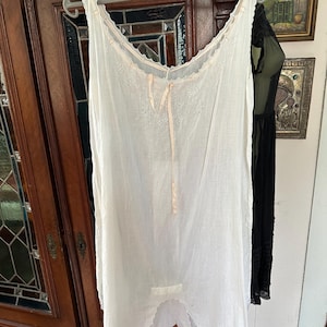 May include: A white vintage slip dress with a scalloped neckline and hem, hanging on a metal hanger. The dress features delicate embroidery and a light pink ribbon detail at the neckline. The dress is made of a lightweight fabric.