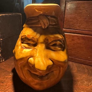 May include: A ceramic pitcher shaped like a smiling face with a brown glaze. The pitcher has a wide brim and a small, round spout. The face has a wide, toothy grin and a prominent nose.