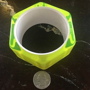May include: A neon yellow, geometric bangle bracelet with a white interior. The bracelet has a square shape with rounded corners and green swirled accents. A U.S. quarter is next to the bracelet for size comparison.