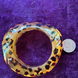 May include: A chunky, oval-shaped bangle bracelet with a tortoiseshell pattern. The bracelet is a warm amber color with dark brown spots. A US quarter is next to the bracelet for size comparison.