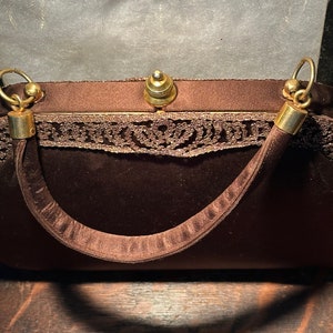 May include: A brown velvet evening bag with a gold clasp and a lace trim. The bag has a single strap and a gold top handle.