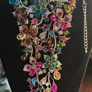 May include: A statement necklace featuring a cascading floral design crafted with multi-colored faceted crystals and rhinestones in shades of pink, blue, green, purple, and amber, set in a gold-tone metal frame with an adjustable chain link closure.