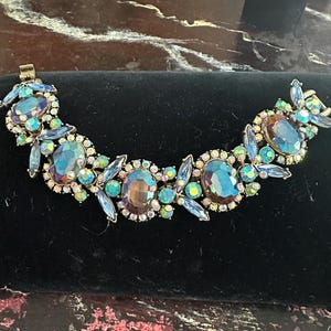 May include: A vintage bracelet with iridescent blue and purple oval and round gemstones. The bracelet has a gold-tone metal setting and features a floral design with smaller blue crystal accents. The bracelet is displayed on a black velvet surface.