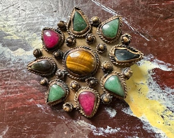 May include: A gold-toned star-shaped brooch with a variety of gemstones, including green, red, and brown. The brooch is decorated with small beads and a rope-like design.