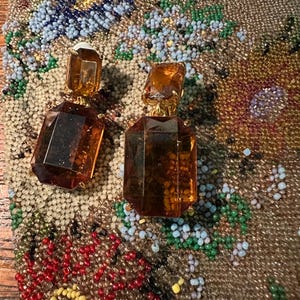 May include: A pair of vintage amber-colored, geometric earrings with gold-tone metal accents. The earrings are set against a backdrop of a beaded, floral-patterned fabric in shades of brown, blue, green, and red.