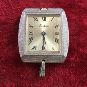 May include: A silver square-shaped pocket watch with a white face and black Roman numeral hour markers. The watch has a small, silver pendant attached to the bottom. The watch face says "Endura" and "Swiss Made".