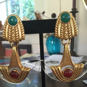 May include: A pair of gold tone dangle earrings with green and red stones. The earrings have a geometric design with a textured finish.